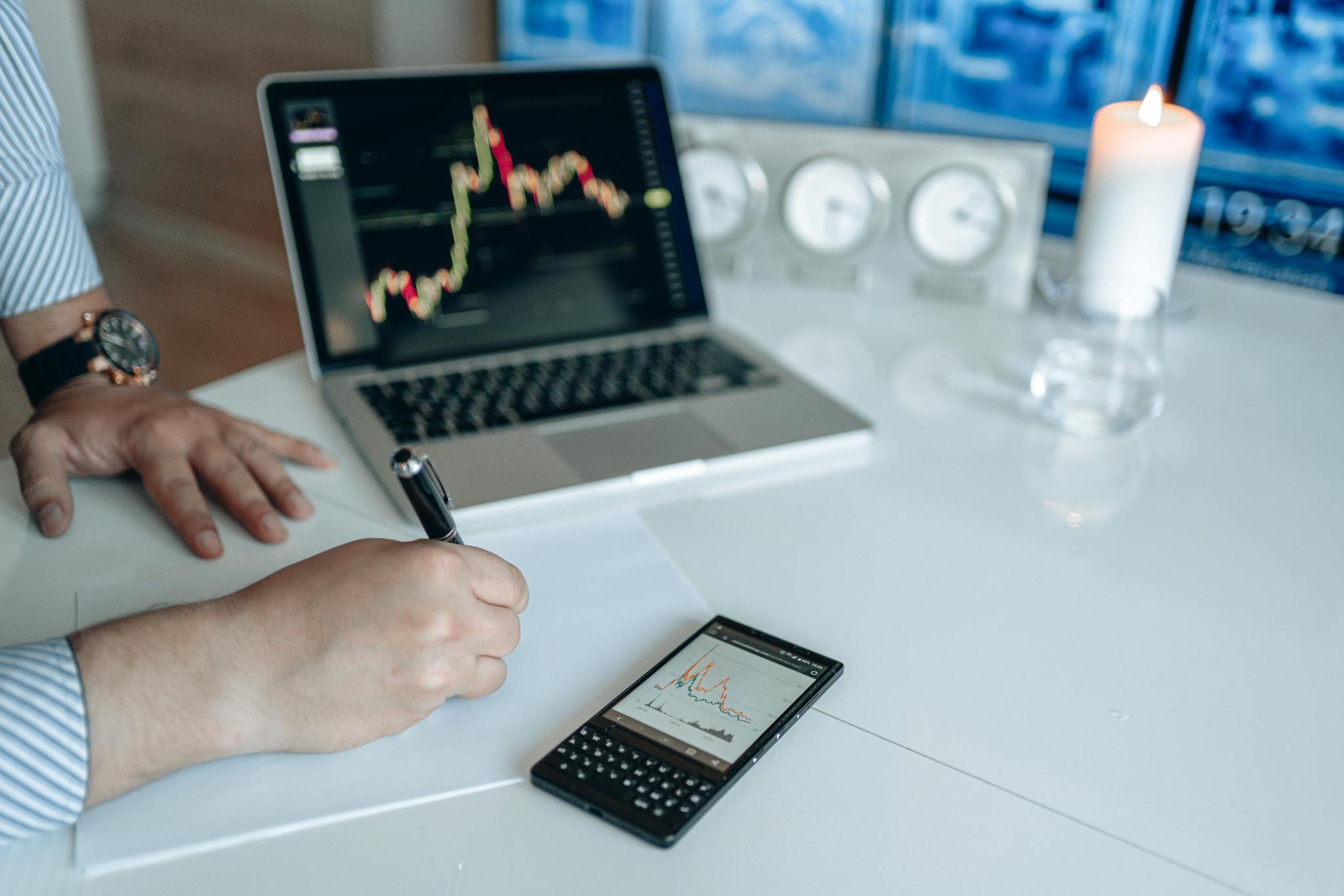 Business analyst writing with laptop and smartphone showing trend graphs on a white desk.