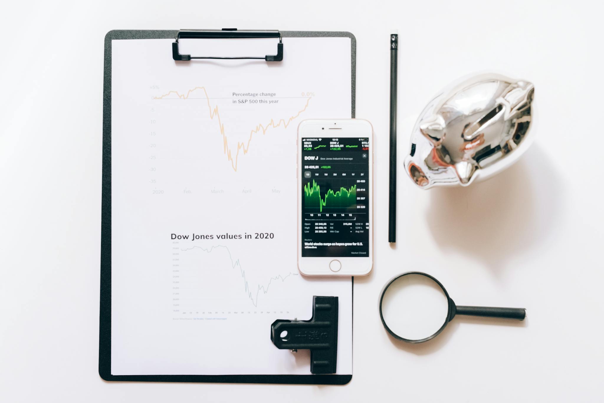 Top view of financial analysis tools including a cellphone, clipboard, magnifying glass, and piggy bank on a white desk.